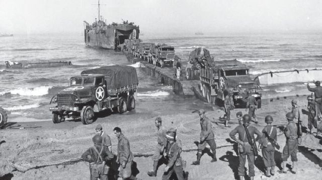 Tropas de EE.UU. desembarcando en Gela. Prisioneros italianos son conducidos por la Policía Militar mientras desembarcan los vehículos del 401º Batallón antiaéreo. 11 de julio de 1943